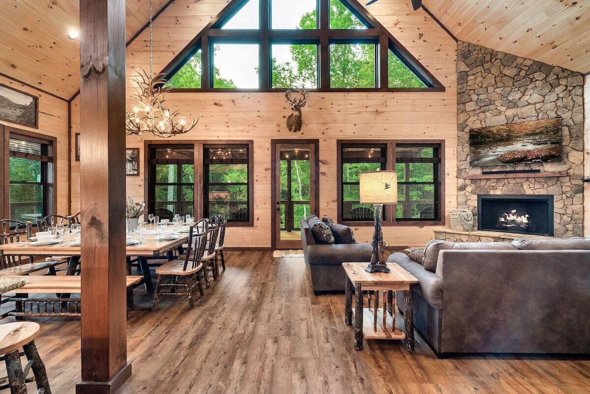 A spacious interior with a high ceiling, wooden floors, a stone fireplace, and large windows offering a view of the outdoors.