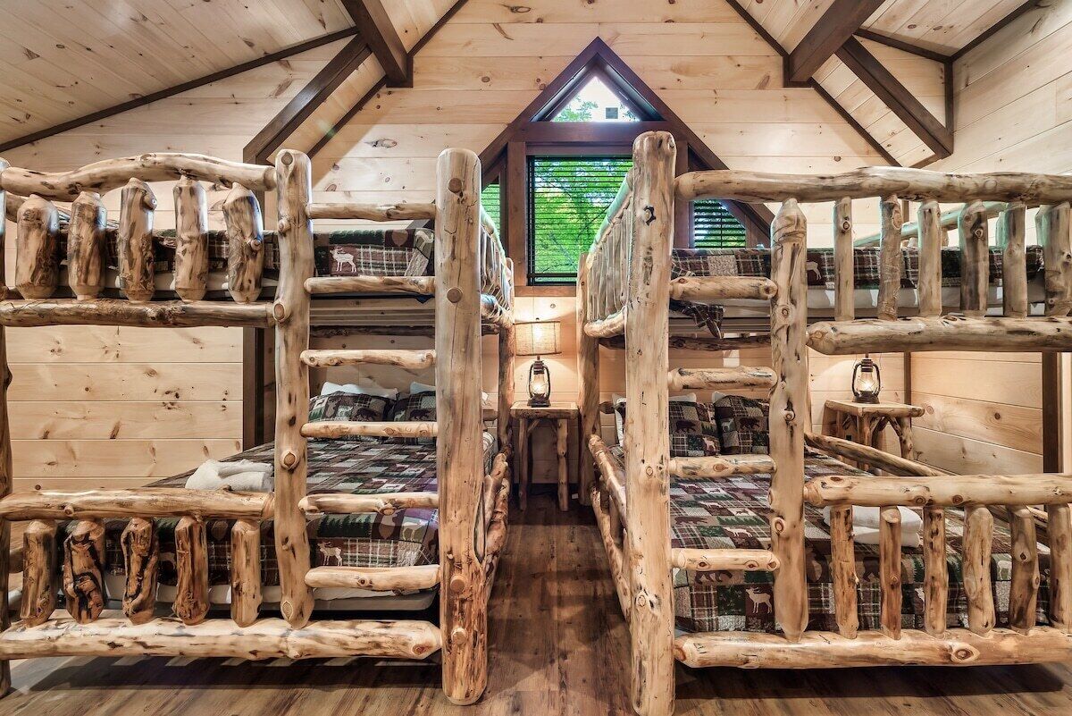 A cabin with wooden bunk beds arranged in a central aisle, a small table with a lantern, and a window with a view of greenery.