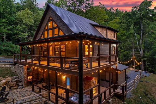 A multi-level wooden cabin with a balcony, stone foundation, and string lights.