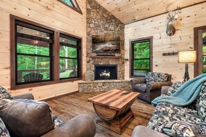 A cozy living room with a stone fireplace, wooden furniture, and large windows offering a view of the forest.