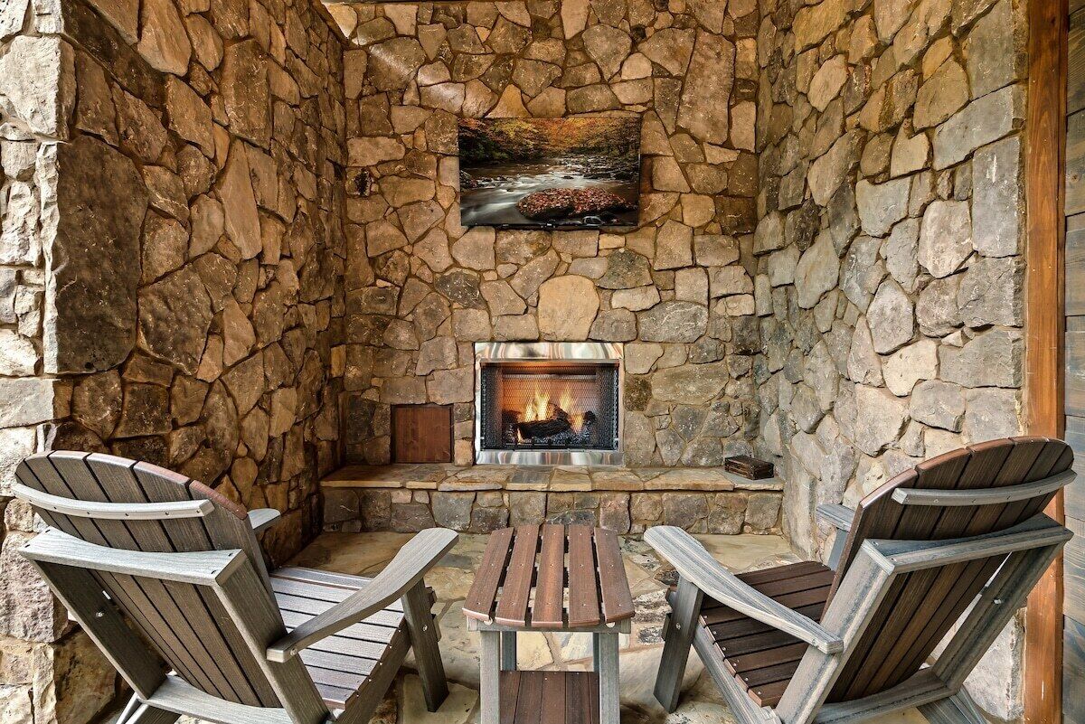 A stone-walled outdoor seating area with a fireplace, two Adirondack chairs, and a small table.