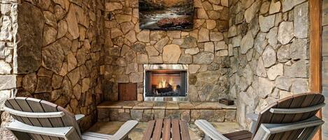 A stone-walled outdoor seating area with a fireplace, two Adirondack chairs, and a small table.