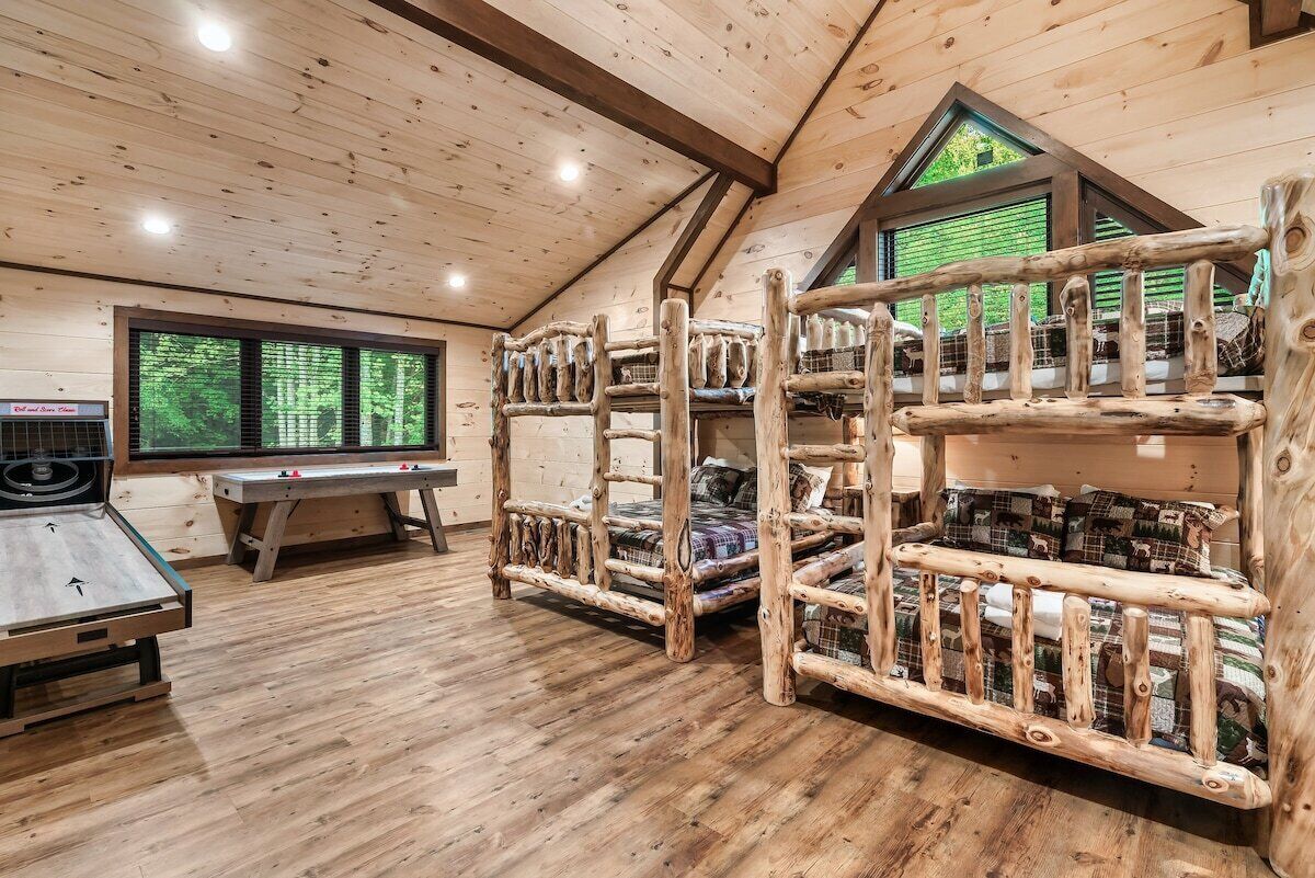 A room with a sloped ceiling, wooden bunk beds, a table, and a window with a view of greenery.