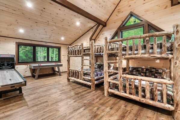 A room with a sloped ceiling, wooden bunk beds, a table, and a window with a view of greenery.