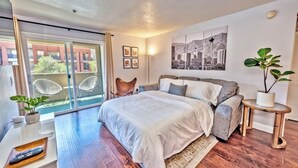 Une chambre moderne avec un grand lit, une porte-fenêtre coulissante donnant sur un balcon, un tableau représentant un paysage urbain et une plante en pot.
