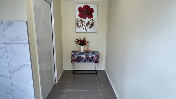 A hallway with a tiled floor, a door, a wall-mounted light fixture, and a table with a vase of flowers. - Brandnew bedroom in a beautiful suburb  (Saint Albans)