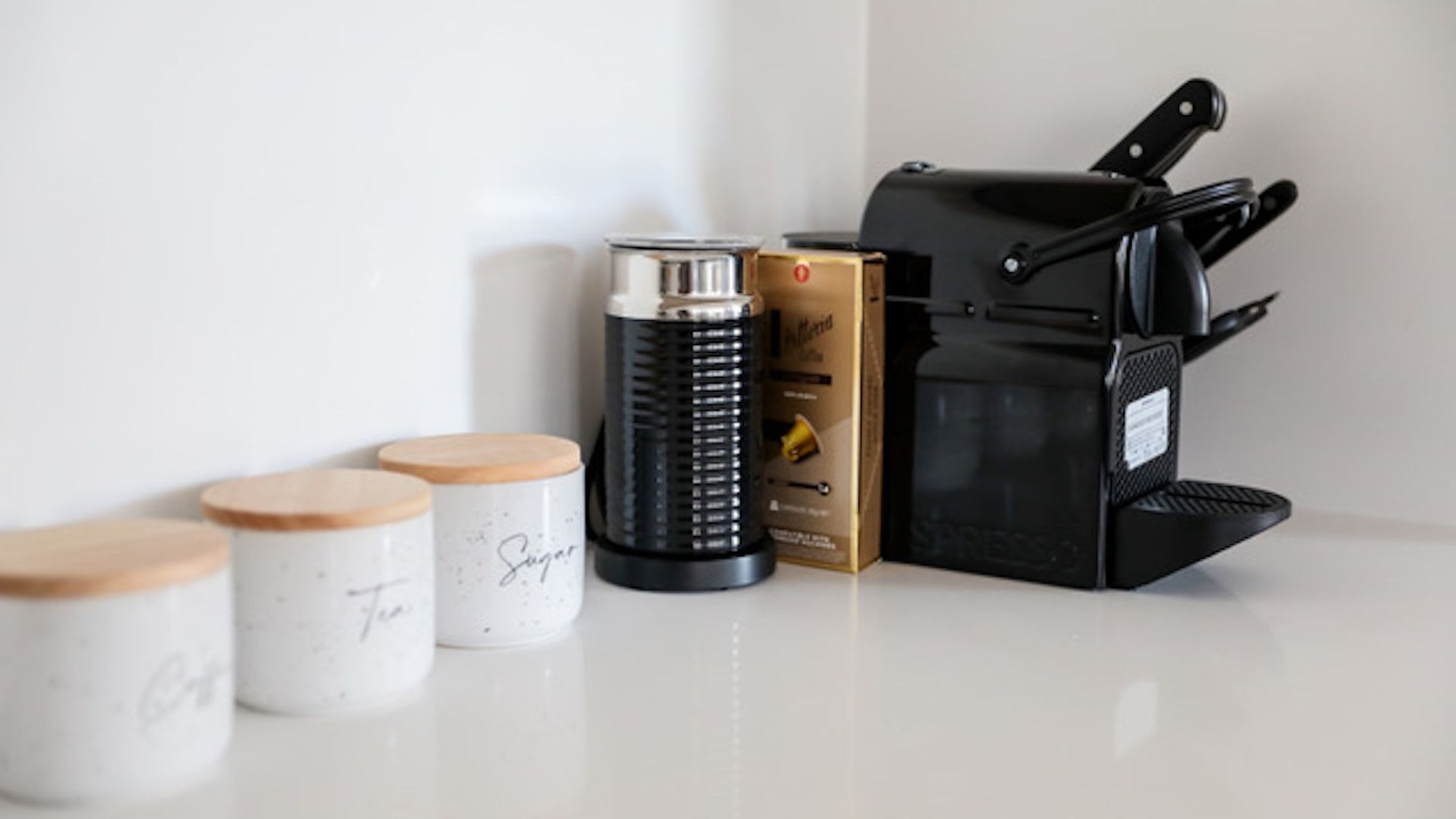 A kitchen counter with three jars labeled "Sugar," "Tea," and "Coffee," a black coffee maker, and a box of coffee pods.