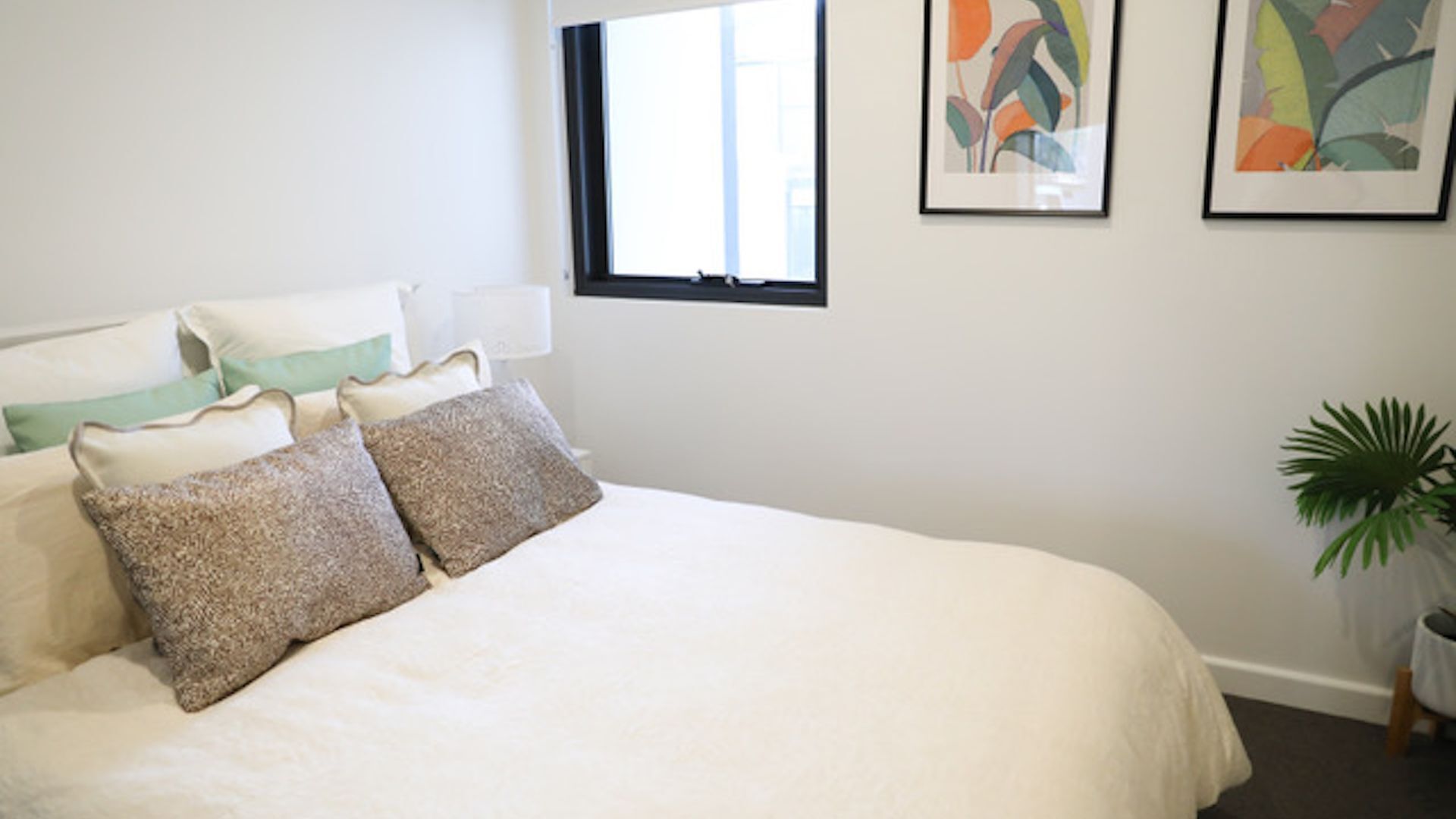 Un dormitorio con una cama grande, varios cojines, una ventana y dos cuadros enmarcados en la pared.