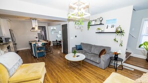 Een moderne woonkamer met een grijze bank, een gele fauteuil en een ronde salontafel. Op de achtergrond is een eethoek met een tafel en stoelen te zien.