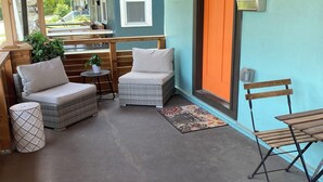 Een overdekte veranda met buitenmeubilair, waaronder een met kussens uitgeruste bank en een rieten stoel, een klein rond tafeltje met een plant, en een metalen bank.