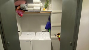 Een wasserij met twee witte voorladers wasmachines en drogers, een plank met opgevouwen handdoeken en een kleine plant, en een blauwe tas hangt aan de muur.