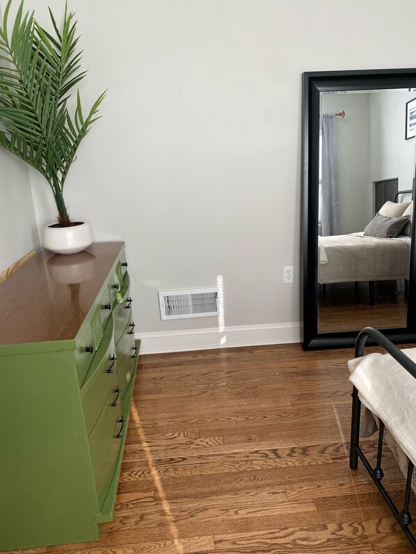 A room with a green dresser, a potted plant, a mirror reflecting a bed, and a vent on the wall.