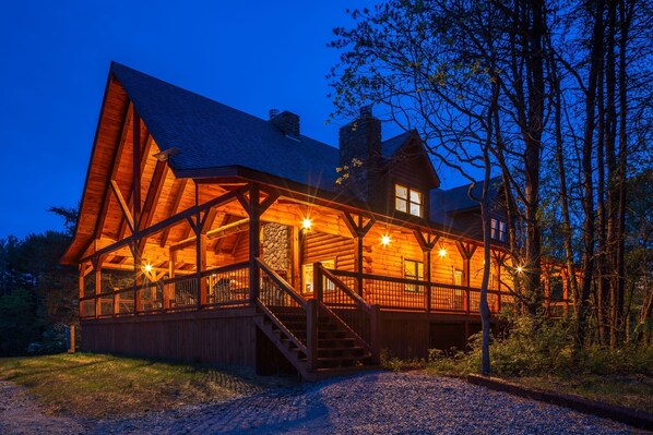 A log cabin with a prominent gabled roof, a stone chimney, and a wooden deck with stairs leading to the entrance.