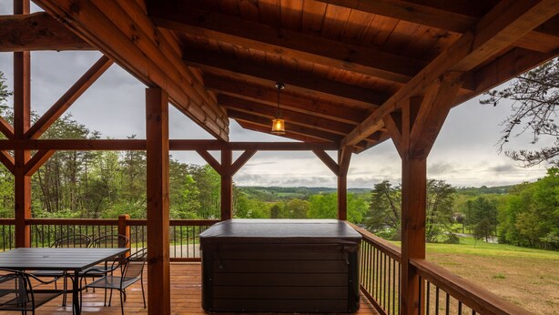 A wooden deck with a table and chairs, a hot tub, and a view of a forested landscape. - Scenic Ridge Lodge - Sleeps up to 16 - w/ Hot Tub (Logan)