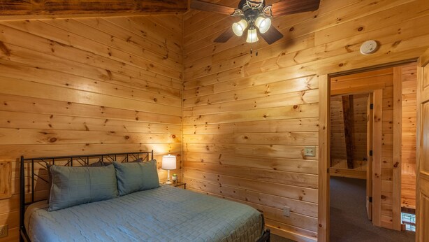 A wooden cabin bedroom with a bed, nightstand, lamp, and ceiling fan.