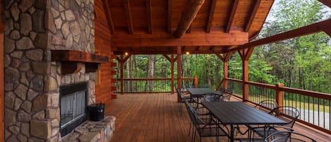 A covered outdoor deck with a stone fireplace, wooden beams, and metal chairs.