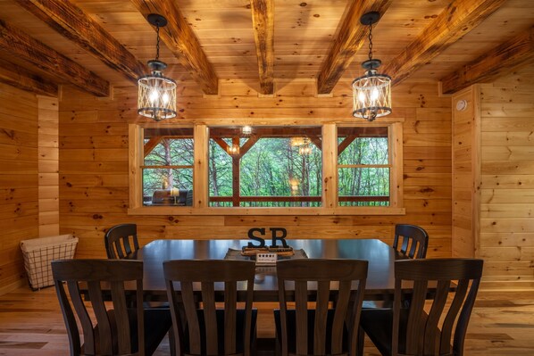 Interior de una cabaña de madera con una mesa de comedor, sillas y dos ventanas que dan a un paisaje de árboles.