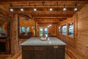 Interior de una cabaña de madera con isla de cocina, comedor y un amplio ventanal con vistas al exterior.