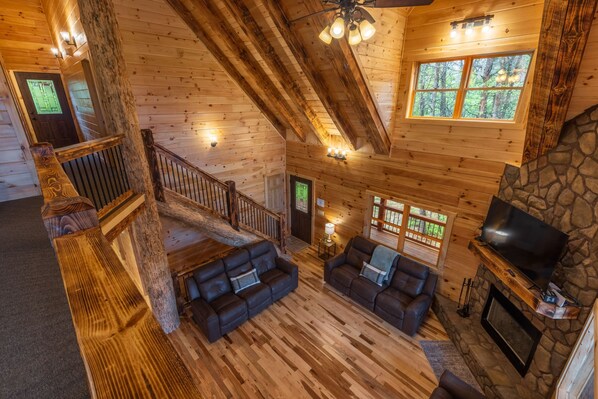 A log cabin interior with a wooden staircase, leather sofas, a stone fireplace, and a television.