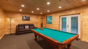 A wooden-paneled room with a pool table, a leather sofa, and a door leading to a screened outdoor area.