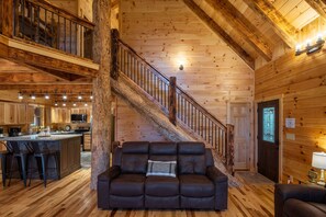 A wooden cabin interior with a central staircase, a living area with a sofa, and a kitchen area with wooden cabinets and a bar.