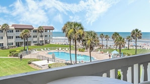 A pool area with a view of the beach and ocean, surrounded by palm trees and a building with balconies.
