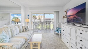 A living room with a sofa, a coffee table, a TV on a cabinet, and a balcony with a view of buildings and palm trees.