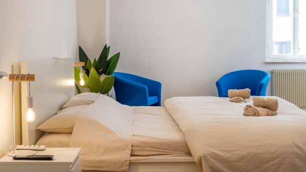 A hotel room with a bed, two blue chairs, a bedside table with a lamp, and a window with a view. - GuestHost - Cividale Medieval Town Apartment (Cividale del Friuli)
