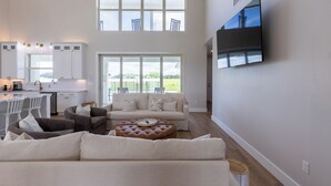 Amplia sala de estar con un ventanal grande, un televisor de pantalla plana montado en la pared y una zona de cocina con armarios blancos y un comedor.