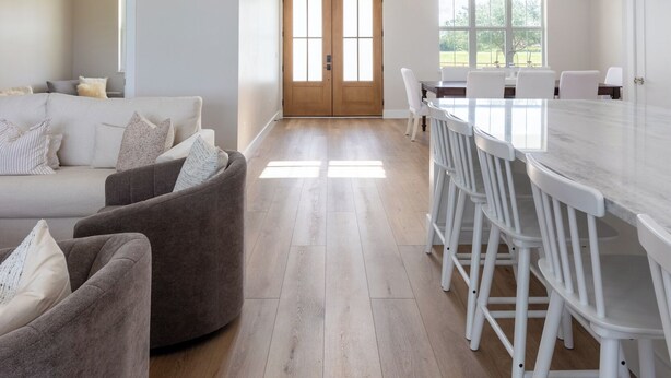 Un salón amplio con un sofá blanco grande y un sillón marrón, un comedor con una mesa y sillas blancas, y una cocina con encimera blanca e iluminación colgante.