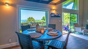 A dining area with a table set for four, large windows offering a view of the ocean, and a balcony with outdoor seating. - Puamana 16B (Princeville)
