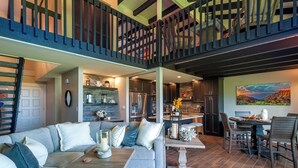 A spacious living area with a high ceiling, a staircase, a kitchen, and a dining area. - Puamana 16B (Princeville)