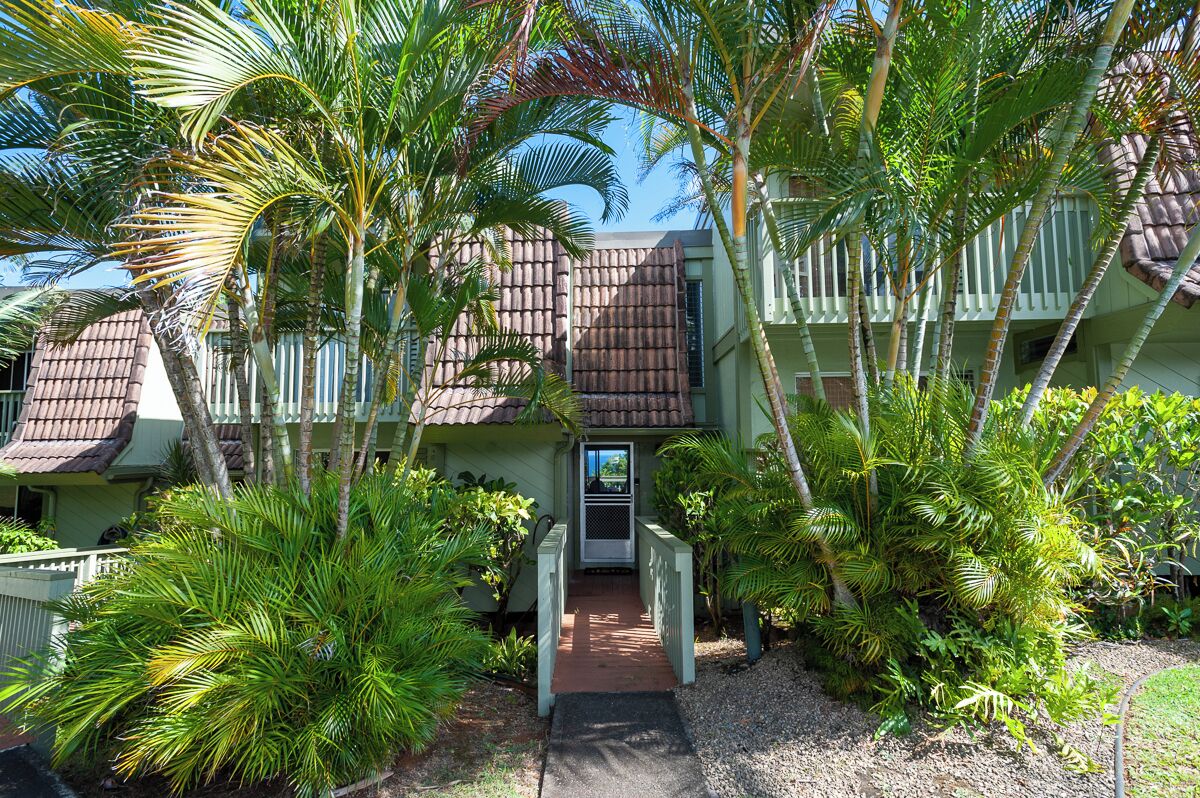 A multi-story building with a green facade and a brown roof, surrounded by lush tropical vegetation.