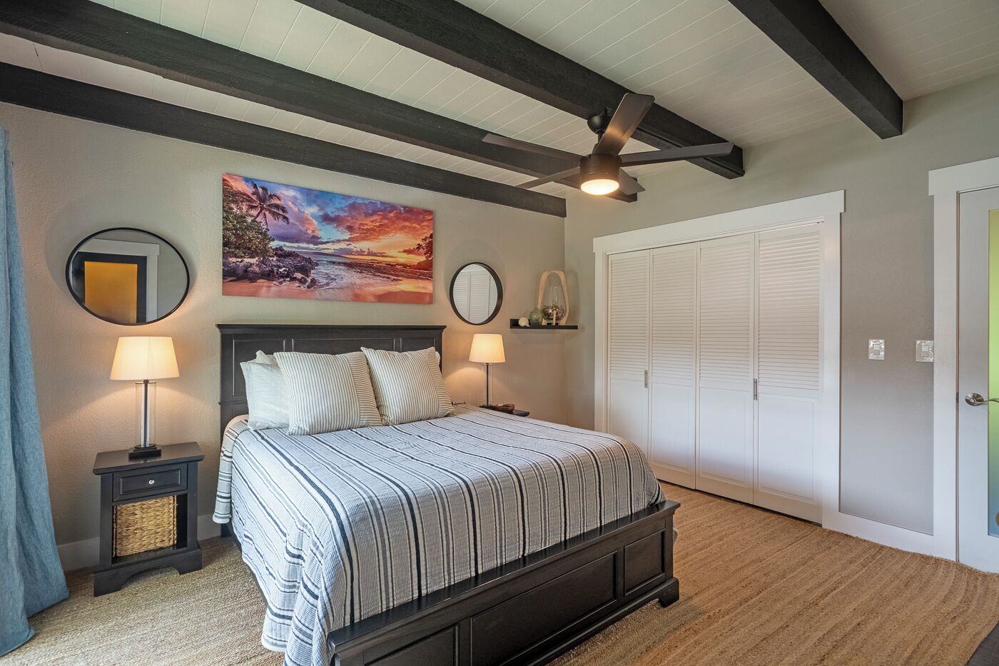 A bedroom with a bed, two bedside tables, a ceiling fan, a large painting, a mirror, and a wardrobe.
