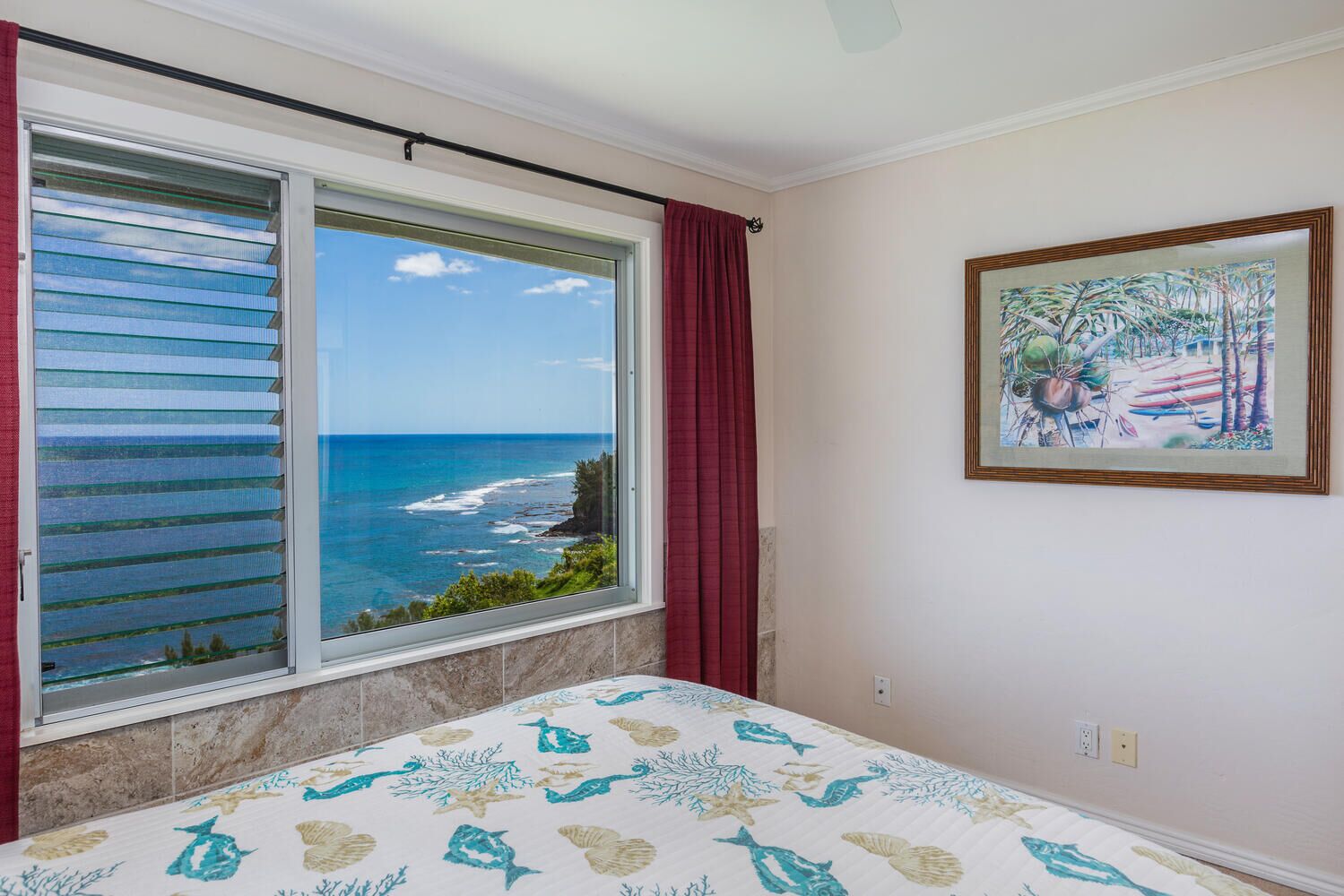 Un dormitorio con un ventanal grande con vistas al océano, una cama con cubrecama de temática marina y un cuadro enmarcado de una escena tropical en la pared.