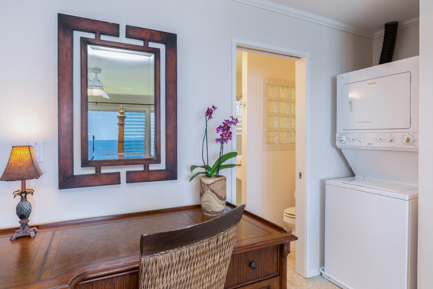 A room with a wooden desk, a lamp, a mirror, a potted orchid, and a washing machine.