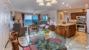 A spacious living area with a glass dining table, comfortable seating, a kitchen with modern appliances, and a view of the ocean through large windows. - Alii Kai 4303 (Princeville)