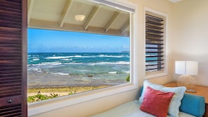 A bedroom with a large window overlooking the ocean, a bed with pillows, a nightstand with a lamp, and a view of the beach and city skyline. - Hanalei Colony Resort F3 (Hanalei)