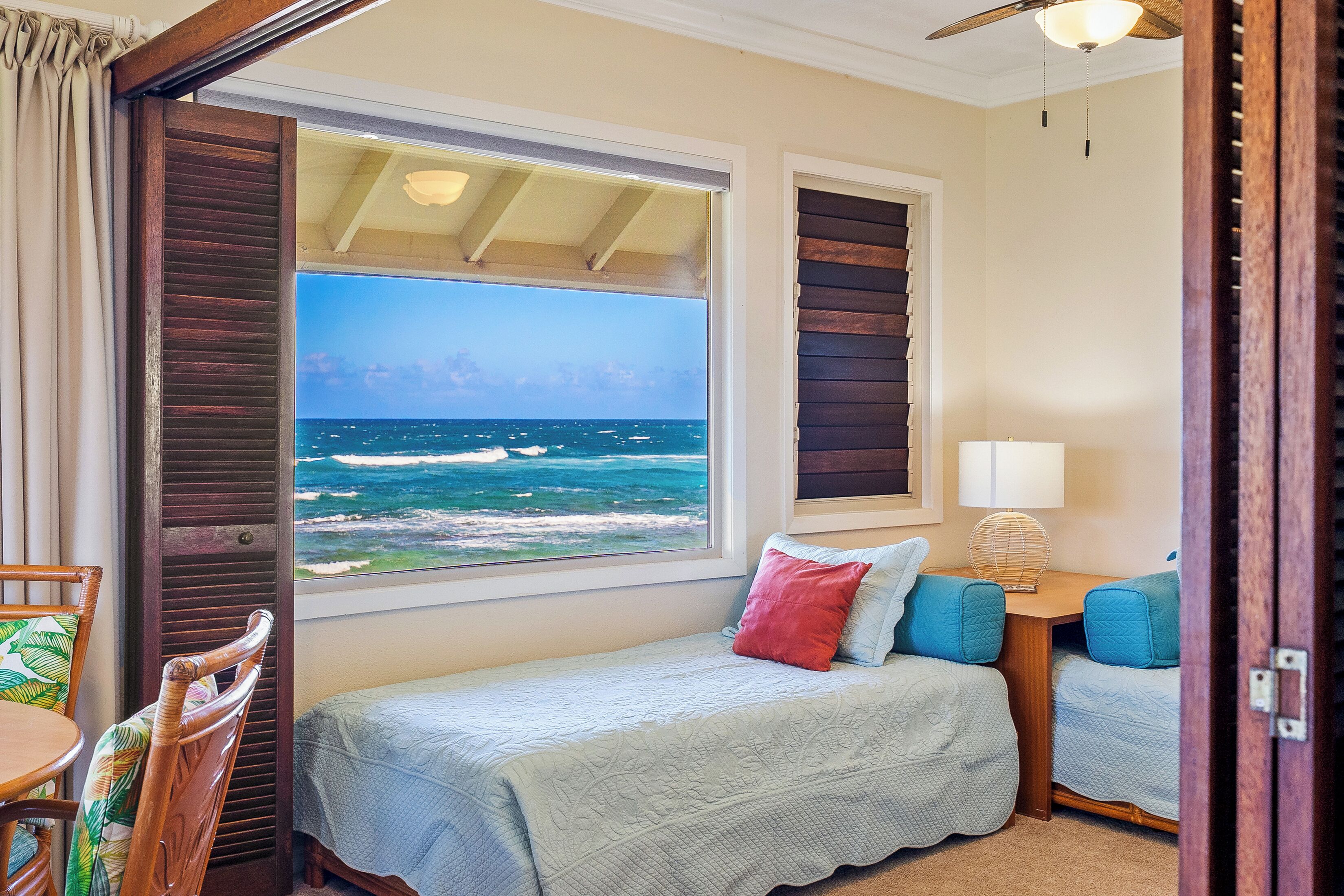 A bedroom with a large window overlooking the ocean, a bed with a light blue bedspread, a red pillow, and a wooden nightstand with a lamp.
