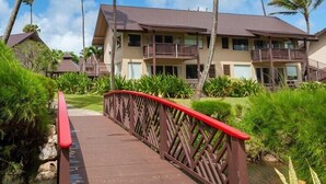A wooden bridge with a red handrail leading to a multi-story building with balconies, surrounded by lush greenery and palm trees. - Hanalei Colony Resort F3 (Hanalei)