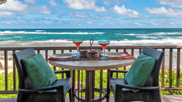 A round table with two wicker chairs, a small tablecloth, and two red drinks. The table is set on a balcony with a view of the ocean. - Hanalei Colony Resort F3 (Hanalei)