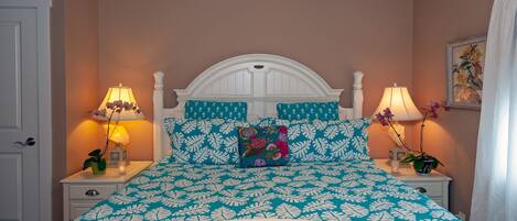 Un dormitorio con cabecera blanca, ropa de cama estampada en verde azulado y dos lámparas de noche, además de una almohada con motivos florales.