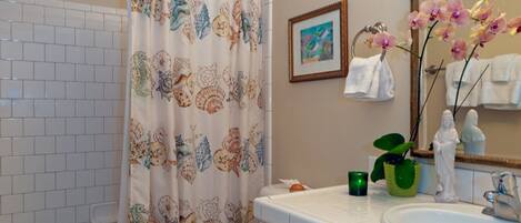 Ein Badezimmer mit Toilette, Waschbecken und Badewanne. Ein Duschvorhang mit Blumenmuster, eine grüne Pflanze auf dem Waschbecken und ein gerahmtes Bild an der Wand.