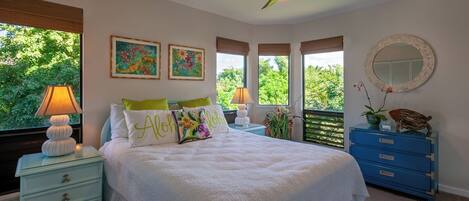Ein Schlafzimmer mit Bett, Nachttischen, Deckenventilator, Kommode und Fenster mit Blick auf Grünflächen.