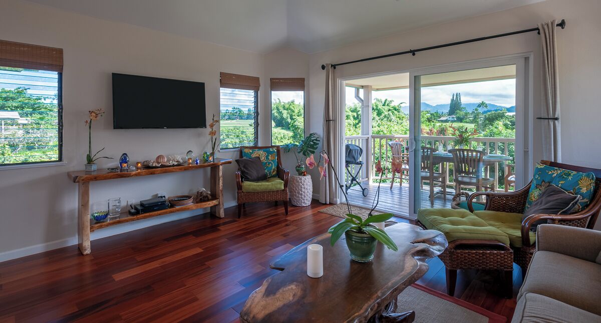 A spacious living room with a wooden floor, a flat-screen TV mounted on the wall, and a balcony with outdoor furniture.