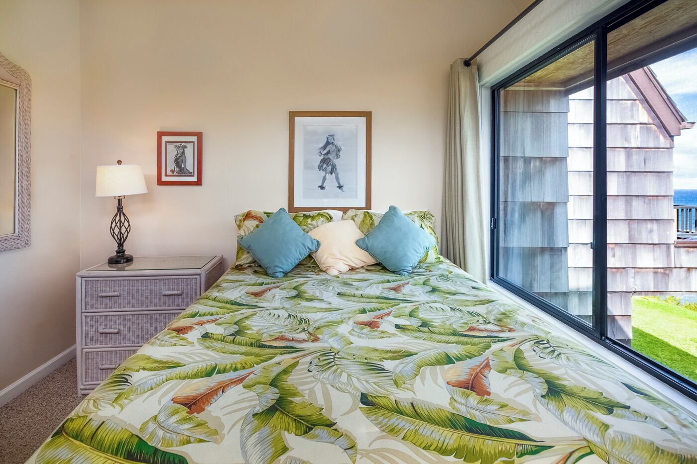 A bedroom with a bed featuring a tropical leaf pattern, a nightstand with a lamp, a framed picture, and a window with a view of a wooden building.