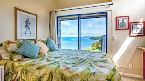 A bedroom with a bed, a large window with a view of the sea, and framed pictures on the wall. - Sealodge J1 (Princeville)