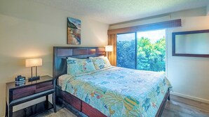 A bedroom with a large bed, a nightstand with a lamp, a painting above the bed, and a window with a view of trees. - Sealodge B4 (Princeville)