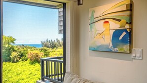 A room with a large window overlooking a lush green landscape and the ocean. There is a painting on the wall and a light switch. - Sealodge B4 (Princeville)