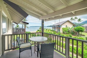 A covered balcony with wicker furniture, a round table, and a view of the ocean and nearby buildings. - Hanalei Colony Resort H4 (Hanalei)